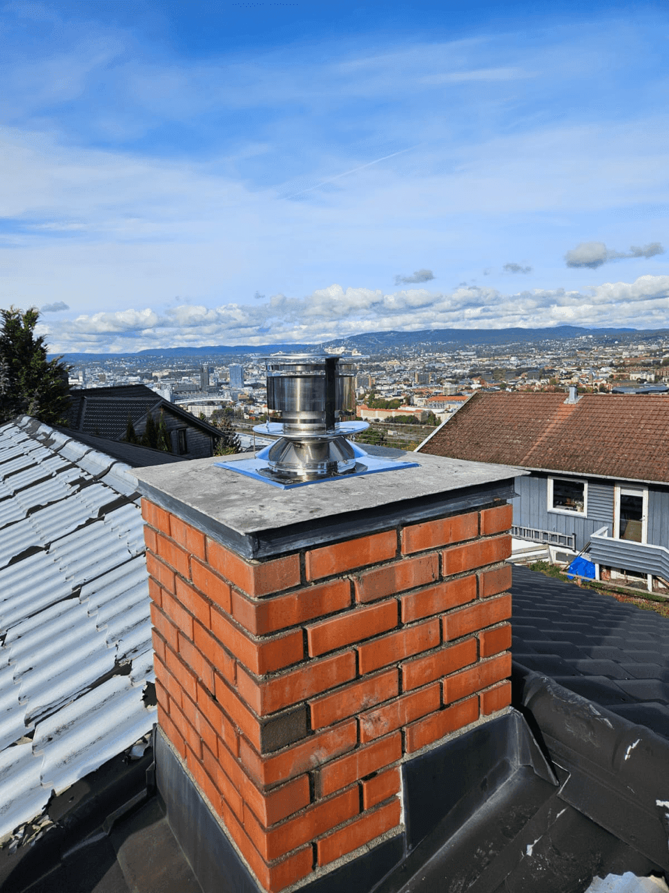 Installasjon av en stålrørshette på toppen av en mursteinskorstein, med verktøy synlig på taket.