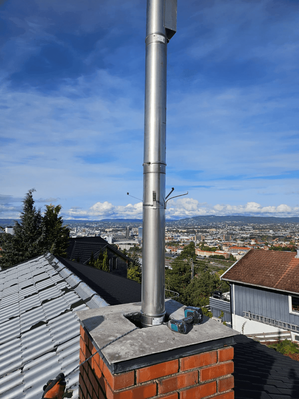 Skorstein etter rehabilitering med ny stålrørshette installert på en mursteinsbase, med utsikt over byen.